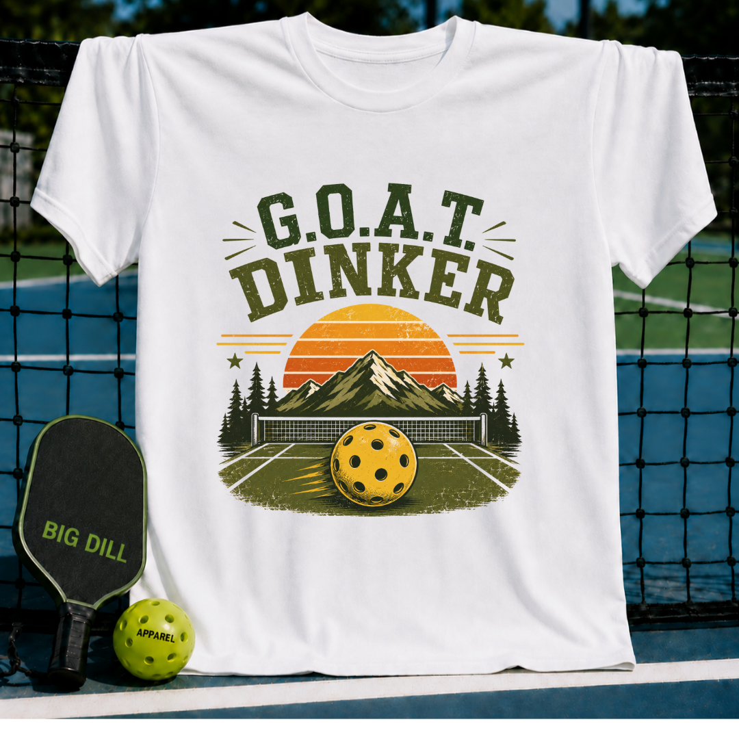 White t-shirt featuring a retro pickleball graphic with mountains, a sunset, and G.O.A.T. DINKER text on a court background.