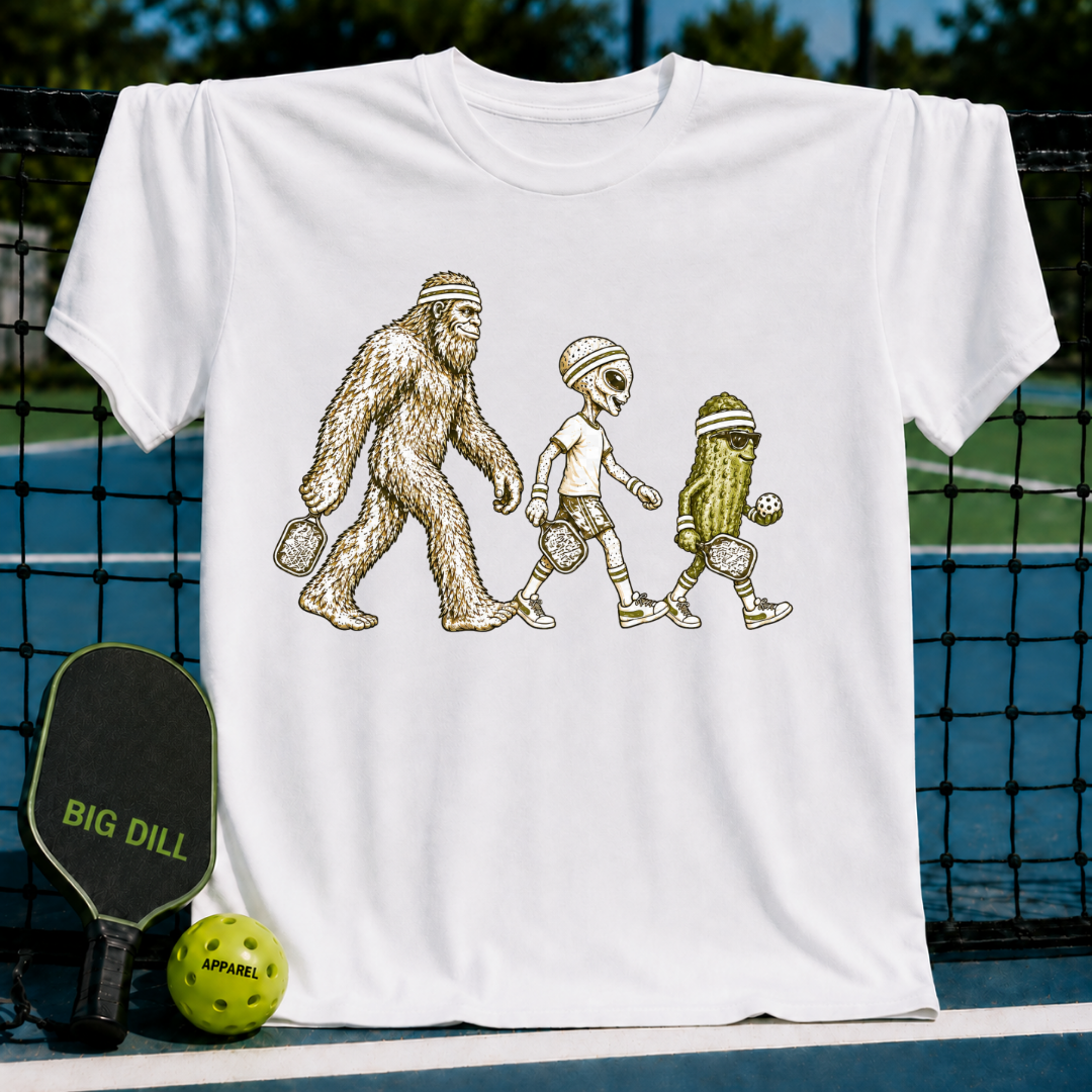 White cotton t-shirt featuring a graphic of Bigfoot, an alien, and a pickle playing pickleball on a court background.