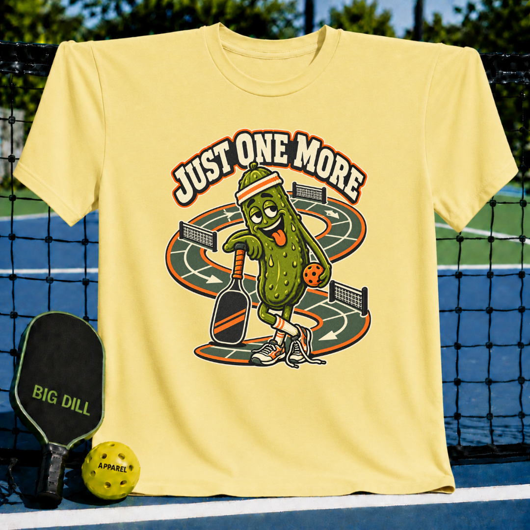 Light yellow crewneck t-shirt featuring a cartoon pickle playing pickleball with the text Just One More.