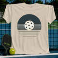 Beige unisex t-shirt featuring a retro charcoal and cream pickleball sunset graphic, displayed on a pickleball court.