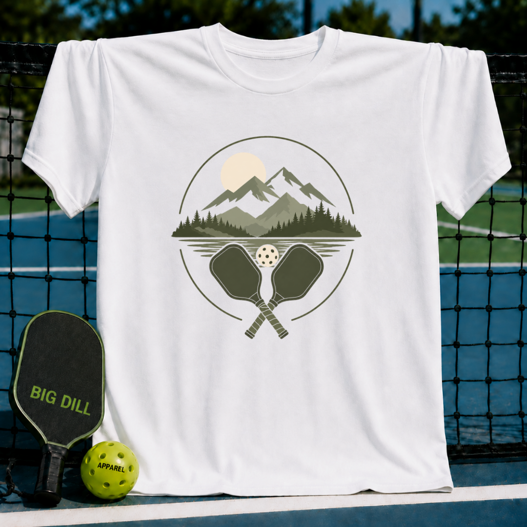 White crew neck t-shirt featuring a mountain, lake, and pickleball paddle graphic on a pickleball court background.