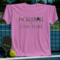 Pink unisex t-shirt featuring Pickleball Couture serif typography, a gold crown, and paddle graphics on a pickleball court.
