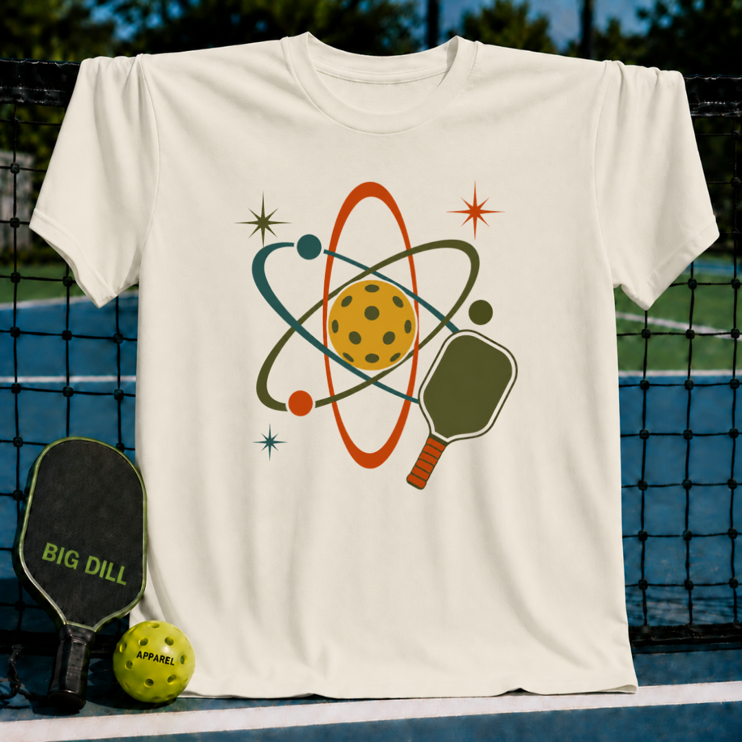 Cream unisex t-shirt featuring a retro mid-century atomic pickleball graphic with a paddle and starbursts on a court background.