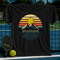Black heavyweight unisex t-shirt featuring a retro circular graphic of mountains, a pickleball net, and a sunset color palette.