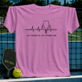 Orchid pink unisex t-shirt featuring a black heartbeat graphic with a pickleball paddle and the text Life Begins At The Kitchen Line.
