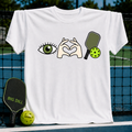 White crewneck t-shirt featuring an eye, heart-shaped hands, and pickleball paddle graphic on a court background.