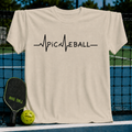 Cream-colored unisex t-shirt featuring a dark brown EKG heartbeat graphic that spells out PICKLEBALL on a pickleball court.