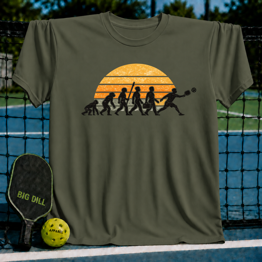 Olive green t-shirt featuring a retro sunset graphic of human evolution ending with a pickleball player lunging for a shot.