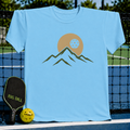 Sky blue unisex t-shirt featuring a minimalist mountain and pickleball eclipse graphic in earth tones.