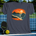 Deep teal unisex t-shirt featuring a vintage oval illustration of a pickleball paddle on a beach at sunset.