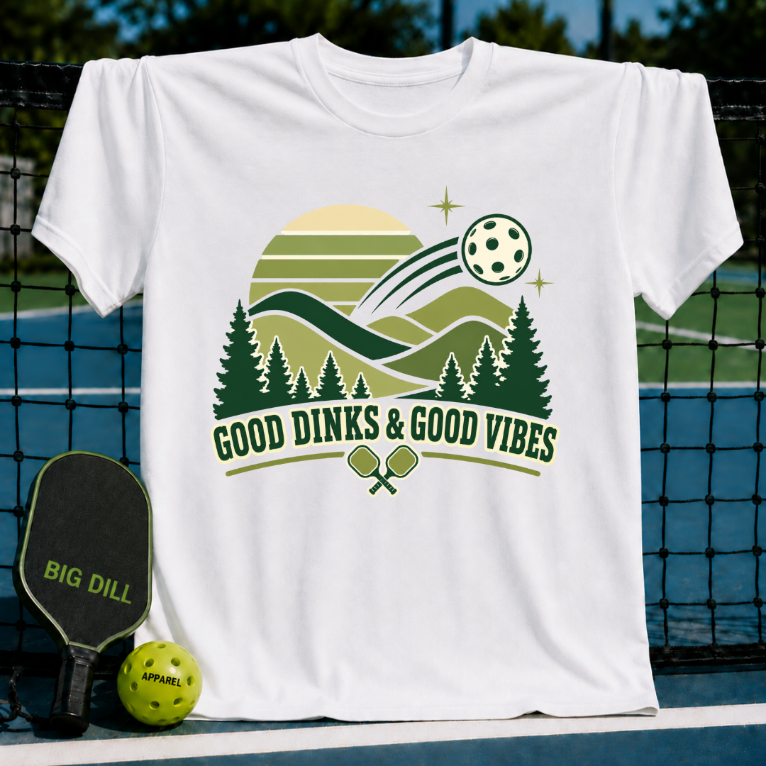 White crewneck t-shirt featuring a green pickleball-themed graphic with pine trees, a sun, and Good Dinks & Good Vibes text.