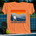 Orange crew neck t-shirt featuring a retro pickleball sunset graphic with the text Play More. Stress Less.