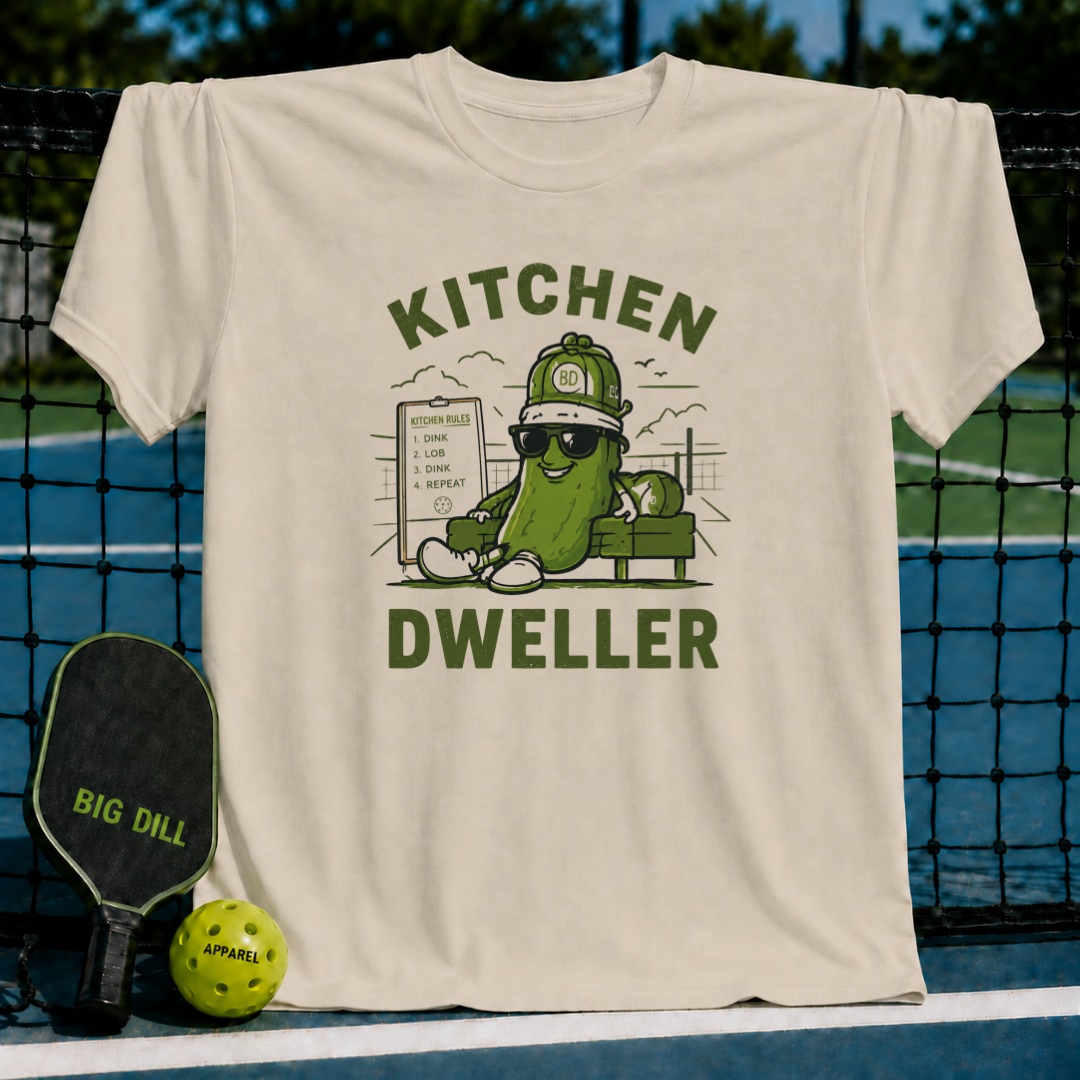 Cream-colored cotton t-shirt featuring a green pickle character wearing sunglasses and a hat with Kitchen Dweller text.