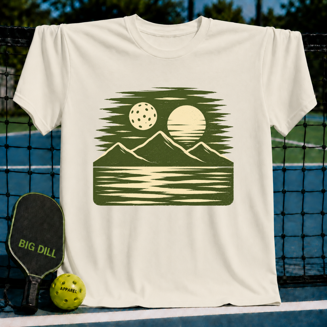 Cream-colored unisex t-shirt featuring a forest green woodblock-style landscape of mountains, a pickleball, and the sun.