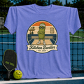 Periwinkle blue unisex t-shirt featuring a retro pickleball graphic of a pickle character at the kitchen line.