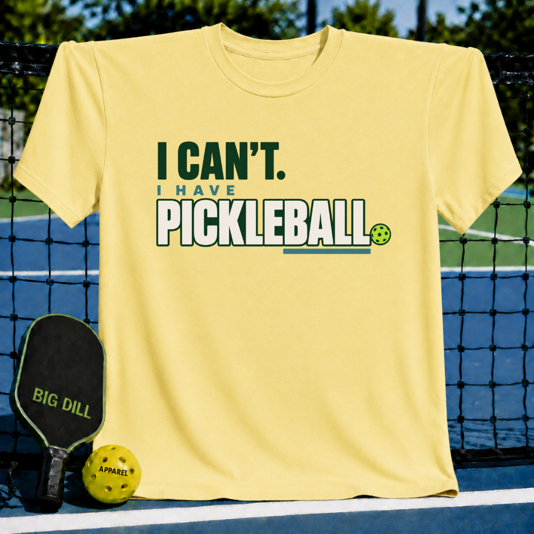 Light yellow crew neck t-shirt featuring I Can't I Have Pickleball text in green and blue, displayed on a pickleball court.