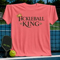 Vibrant coral unisex t-shirt featuring a gold crown and dark olive Pickleball King text on a pickleball court.