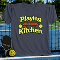 Charcoal grey crewneck T-shirt with yellow and red Playing in the Kitchen pickleball graphic on a court background.