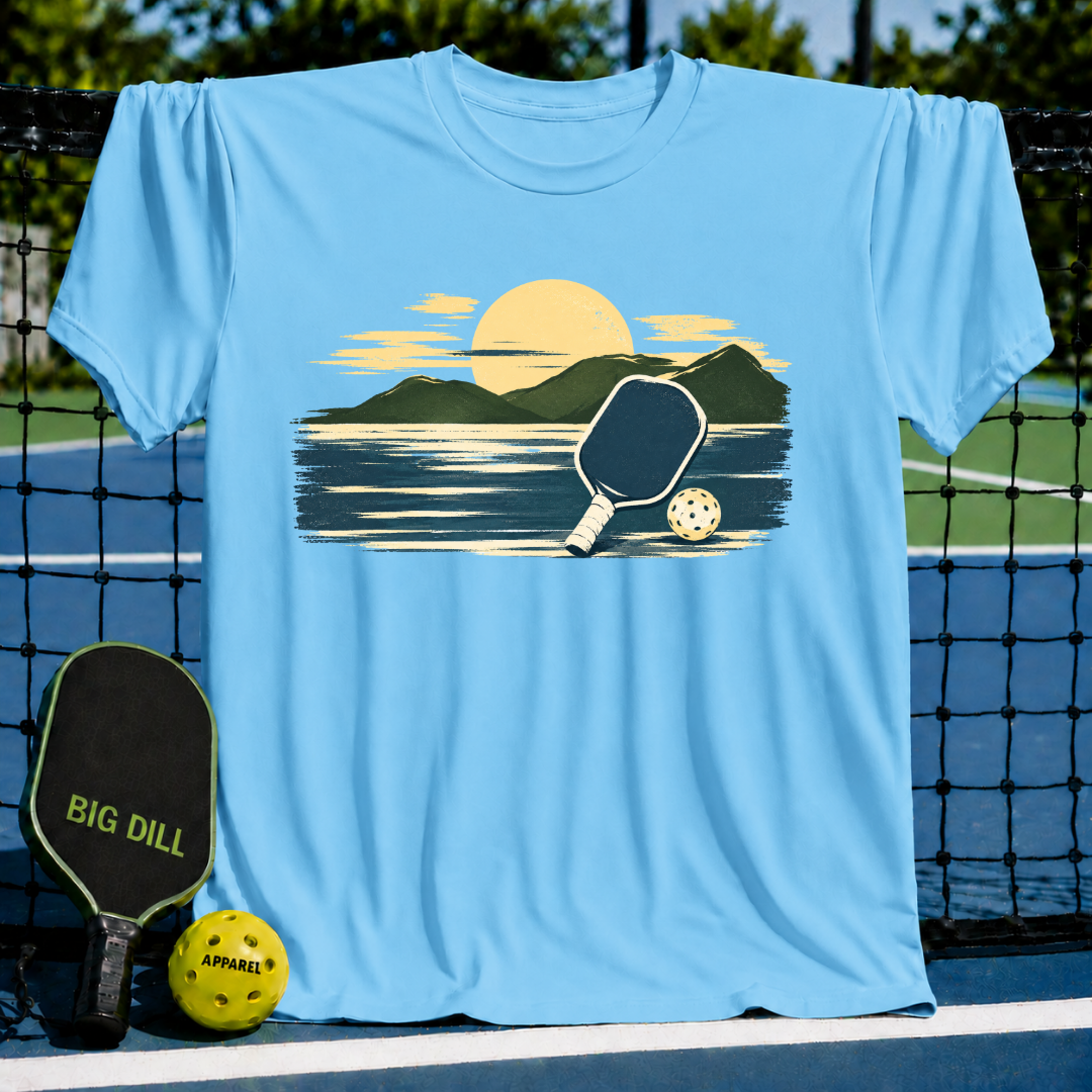 Light blue unisex t-shirt featuring a painterly mountain, paddle, and ball graphic, displayed on a pickleball court.