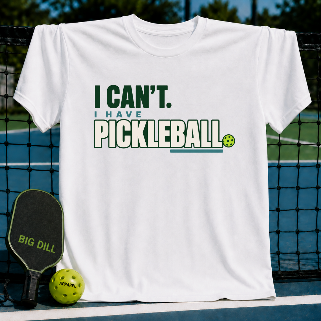 White crewneck t-shirt featuring green I Can't I Have Pickleball text, displayed on a pickleball court net.