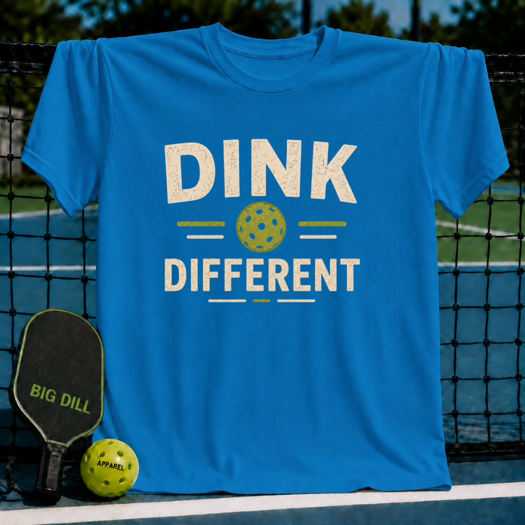 Bright blue Dink Different pickleball t-shirt with a distressed graphic, displayed on a court net with a paddle and ball.