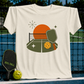Cream-colored crewneck t-shirt featuring a midcentury modern pickleball court graphic with a sun, paddle, and ball.