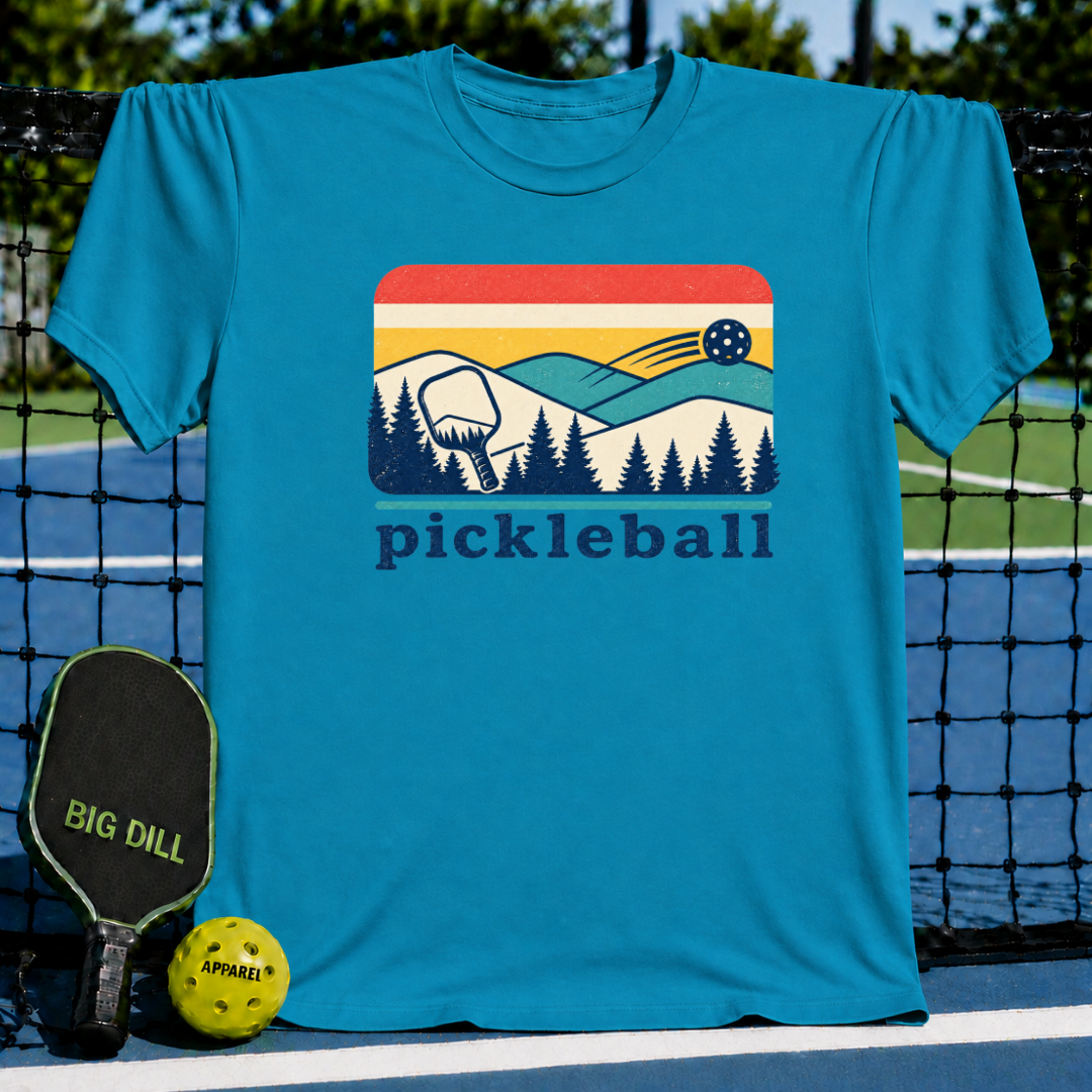 Blue unisex t-shirt featuring a retro mountain, pine tree, and pickleball graphic on a pickleball court background.