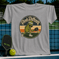 Heather gray unisex t-shirt featuring a retro pickleball graphic of a pickle in a visor hitting a ball on a court.