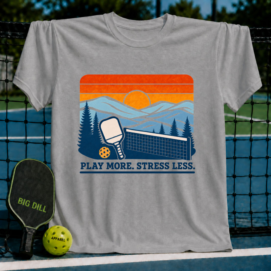Grey t-shirt featuring a retro sunset pickleball graphic with a paddle, net, and the text Play More. Stress Less.