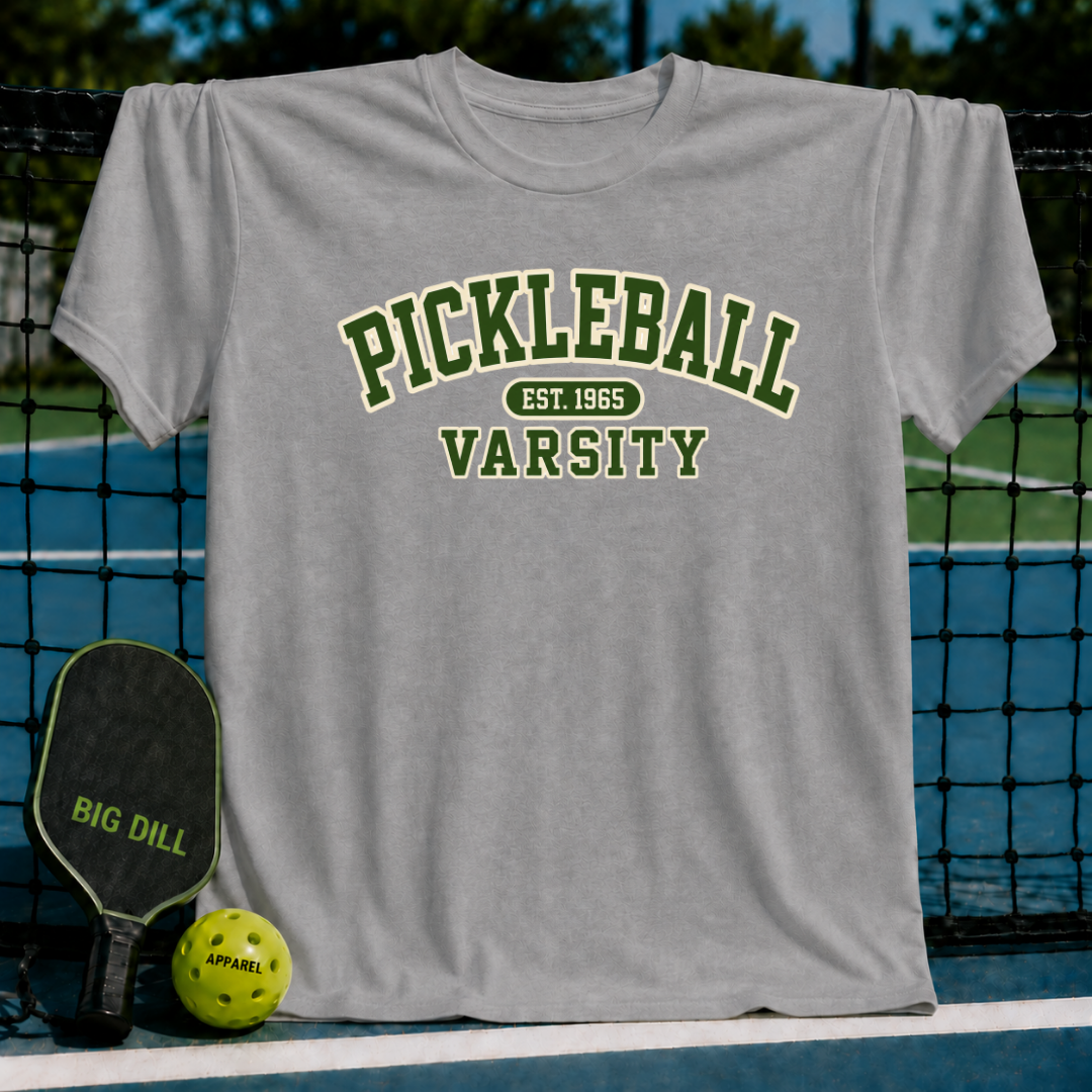 Grey crewneck t-shirt featuring green Pickleball Varsity text, displayed on a pickleball court with a paddle and ball.