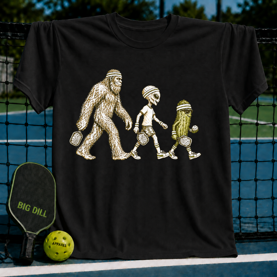 Black cotton t-shirt featuring a graphic of Bigfoot, an alien, and a pickle playing pickleball on a court background.