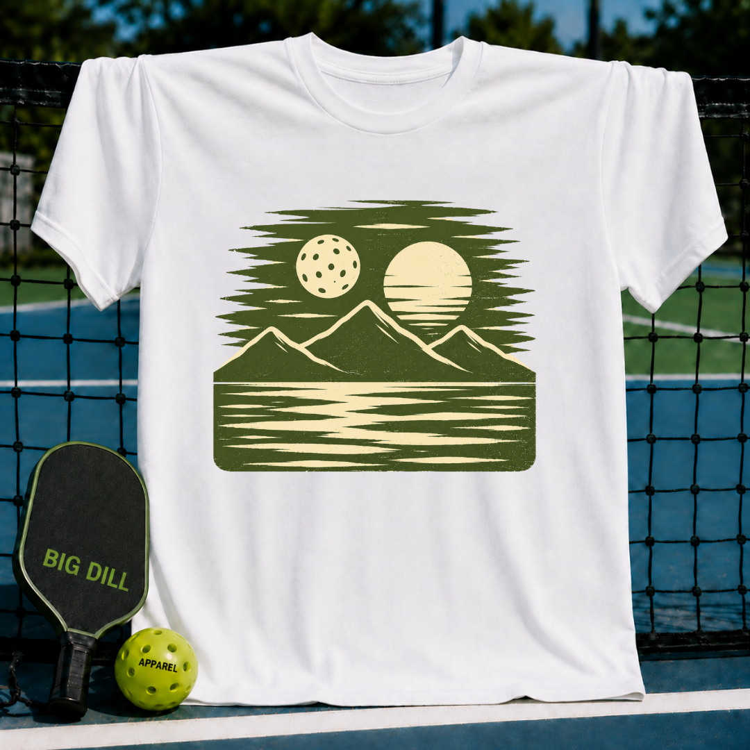 Cream unisex t-shirt featuring a forest green woodblock-style graphic of a pickleball, sun, mountains, and water.