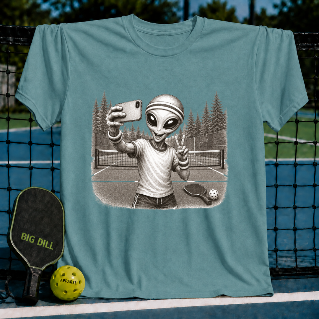 Sage green unisex t-shirt featuring a vintage-style illustration of an alien taking a selfie on a pickleball court.