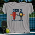 Grey unisex t-shirt with a vintage Gen X Still Dinkin' pickleball graphic, displayed on a court net.