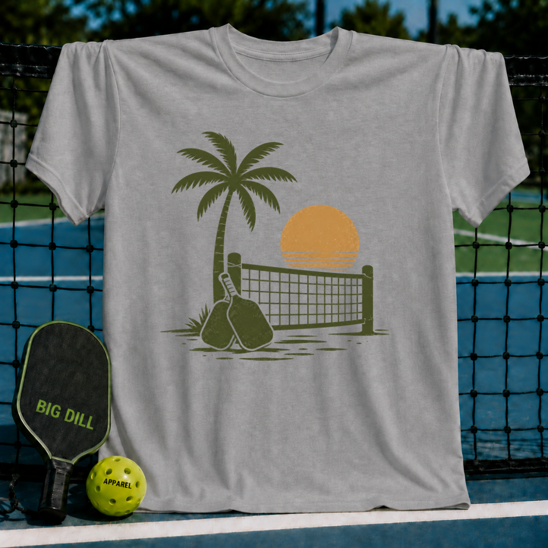 Light gray unisex t-shirt featuring a retro palm tree, pickleball net, and paddle graphic on a pickleball court background.
