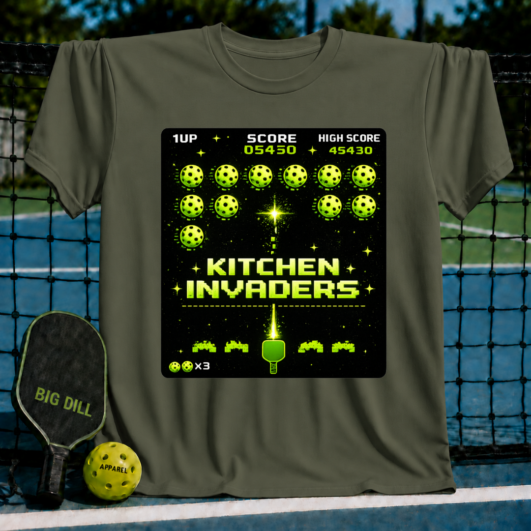 Olive green unisex t-shirt featuring a pixel-art Kitchen Invaders graphic with pickleballs and a paddle on a court background.