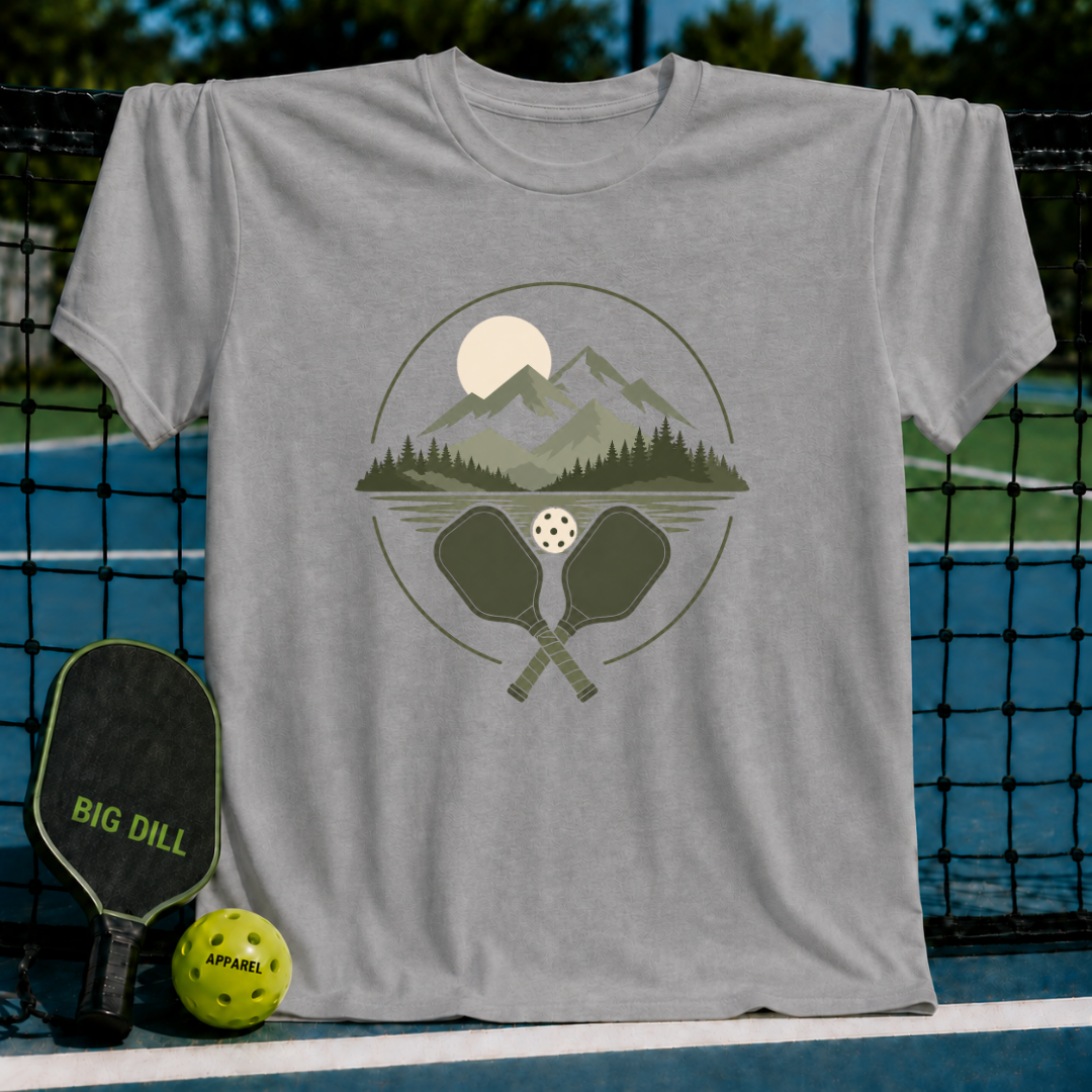 Grey crewneck t-shirt featuring a mountain, forest, and pickleball paddle graphic hanging on a court net.
