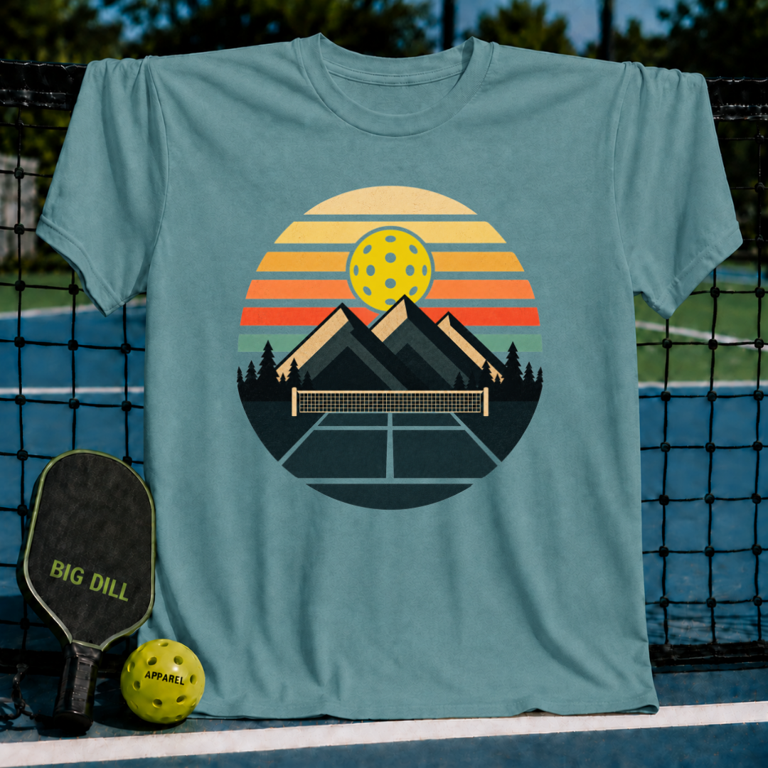 Blue-grey unisex t-shirt featuring a retro circular graphic of mountains, a pickleball net, and a sunset-colored sky.