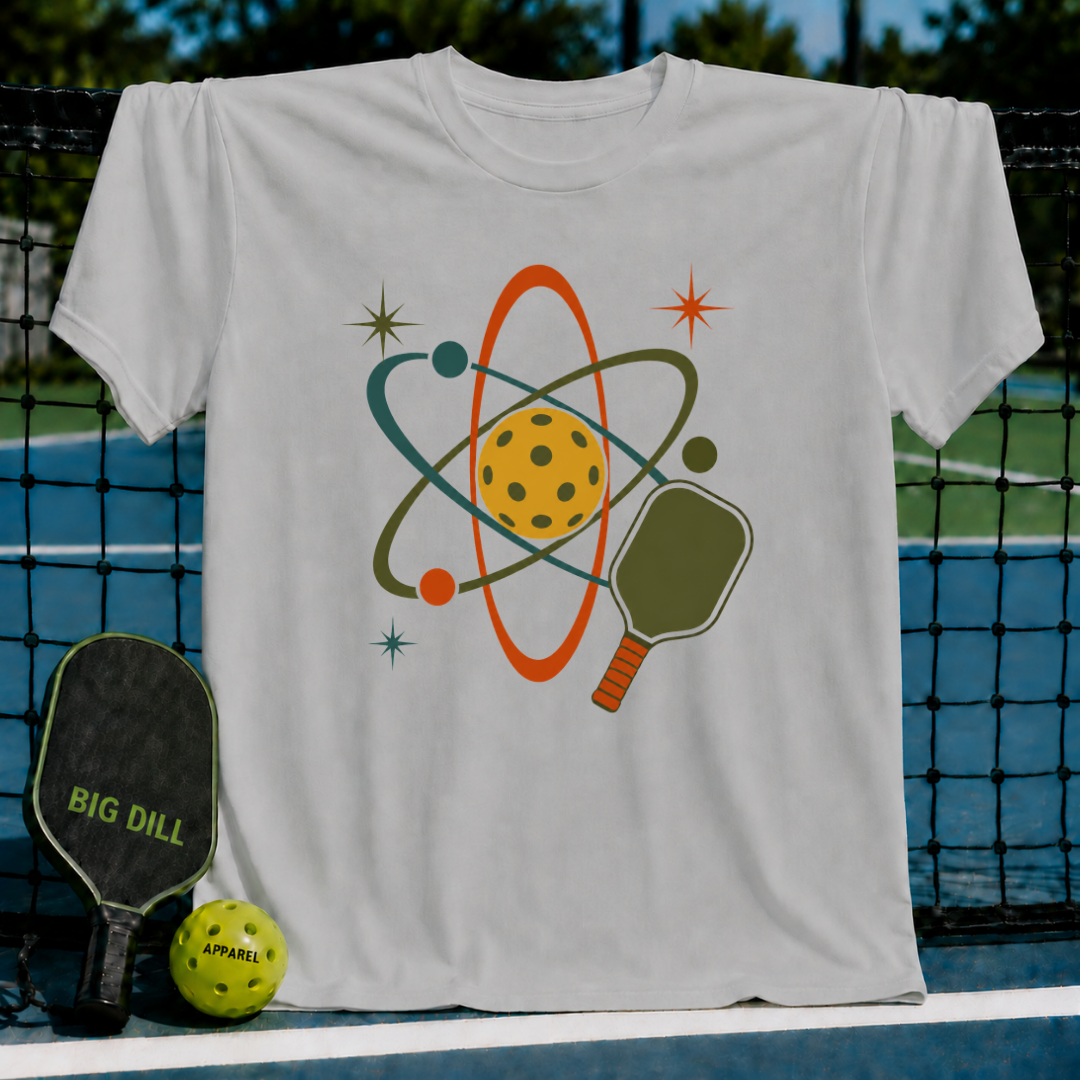 Cream unisex t-shirt featuring a retro mid-century atomic pickleball graphic with a paddle, orbiting rings, and starbursts.