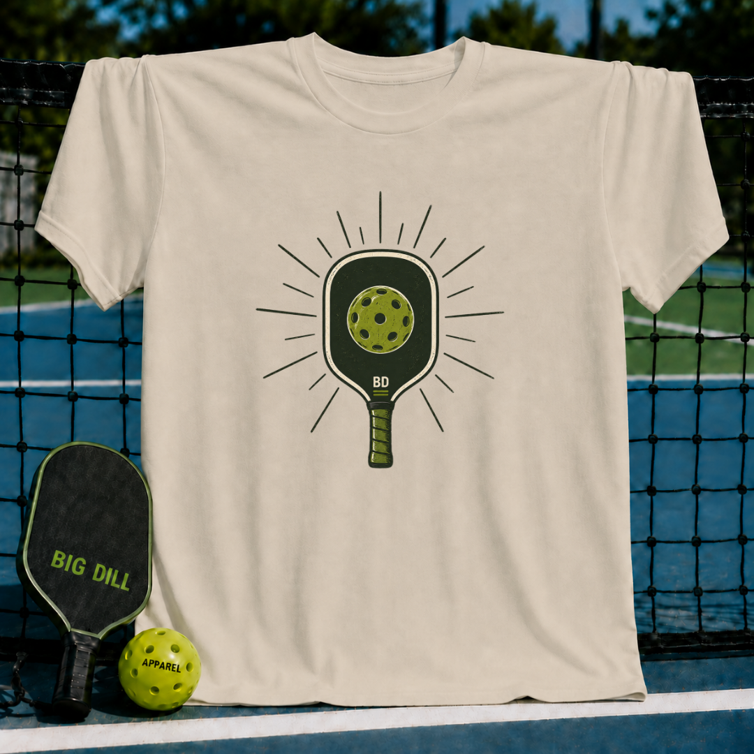 Beige crewneck t-shirt featuring a green pickleball paddle and ball graphic on a pickleball court background.