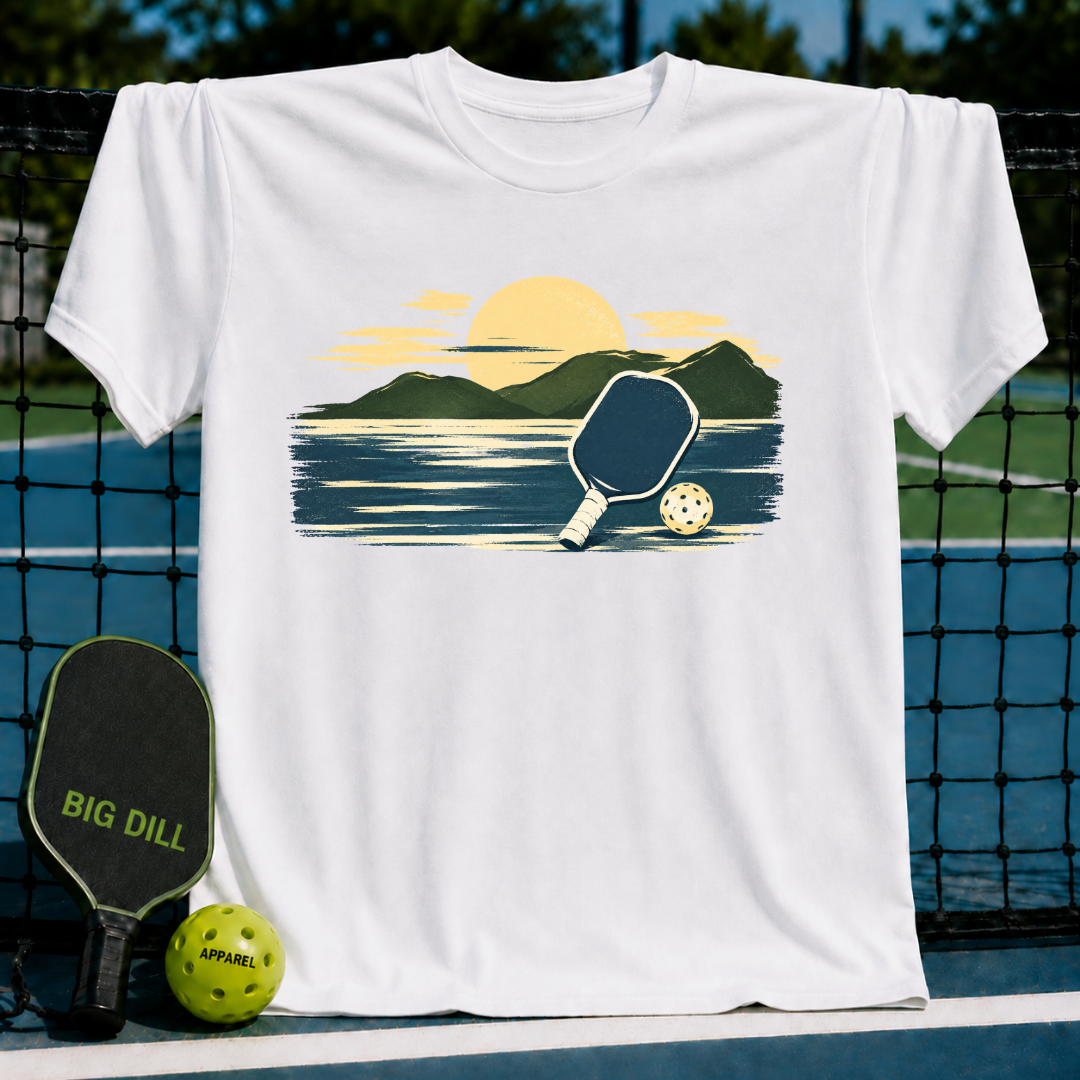 White unisex t-shirt featuring a painterly mountain and pickleball graphic in golden and navy tones on a court background.