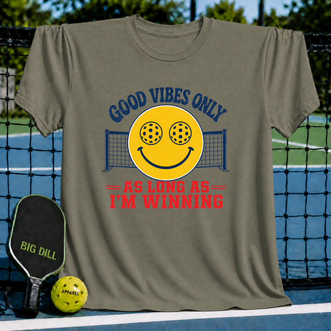 Olive green unisex t-shirt featuring a yellow smiley face with pickleball eyes and a net graphic, with bold text on a court.