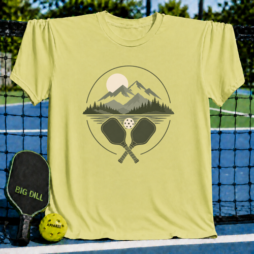 Light yellow crew neck t-shirt featuring a mountain, forest, and pickleball paddle graphic on a pickleball court background.