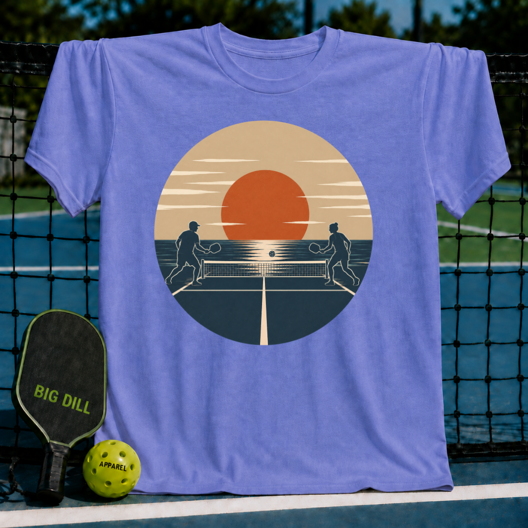 Periwinkle blue unisex t-shirt featuring a retro sunset pickleball silhouette graphic, displayed on a court.