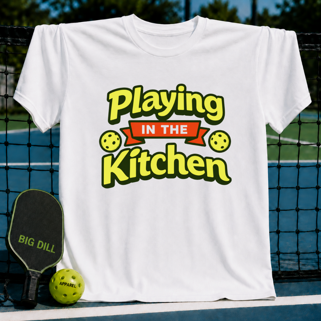White crewneck t-shirt featuring a yellow and red Playing in the Kitchen pickleball graphic, displayed on a court net.