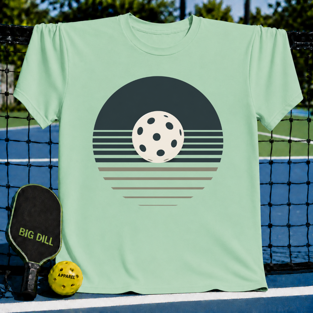 Light green unisex t-shirt featuring a retro pickleball sunset graphic in charcoal and cream on a pickleball court.