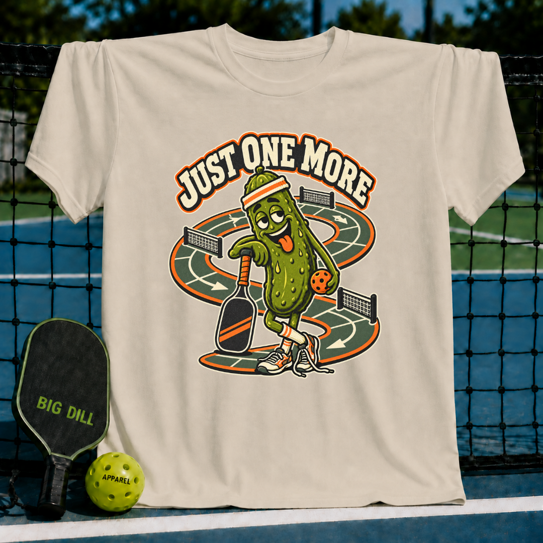 Cream-colored t-shirt featuring a cartoon pickle character playing pickleball with the text Just One More on a court background.