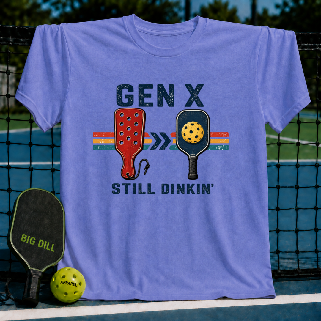 Periwinkle blue t-shirt featuring a retro Gen X Still Dinkin' pickleball graphic, displayed on a court net.