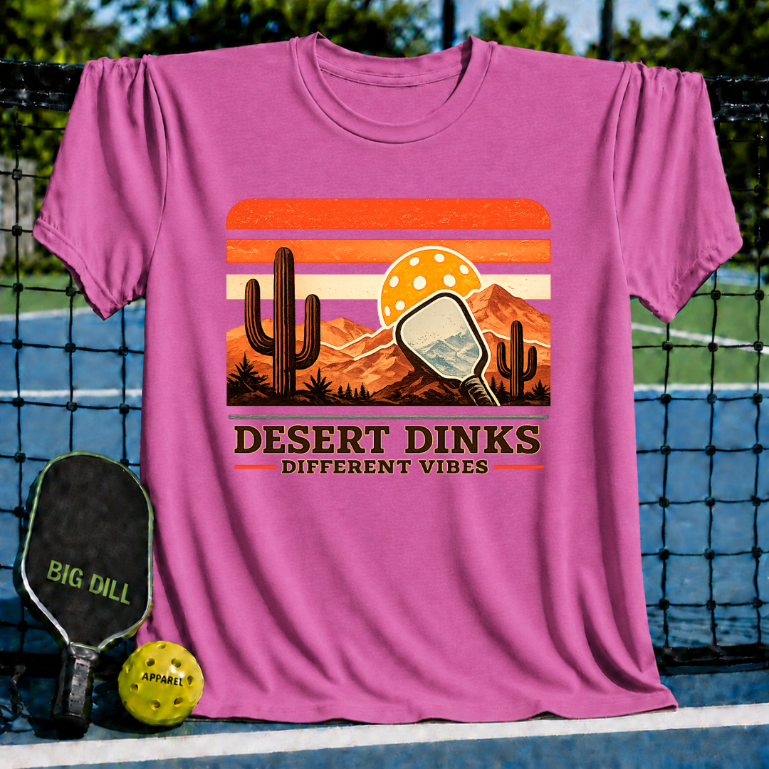 Pink Desert Dinks t-shirt featuring a retro sunset, cactus, and pickleball paddle graphic on a pickleball court.