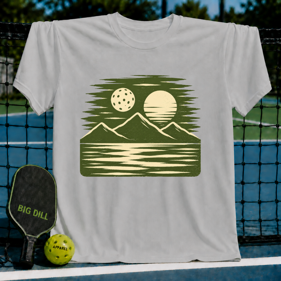 Cream unisex t-shirt featuring a forest green woodblock-style print of a pickleball, sun, mountains, and water reflection.